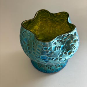 May include: A small glass bowl with an iridescent blue and green finish. The exterior features a raised, textured pattern of circular and organic shapes. The rim is wavy and uneven, while the interior displays a smooth, reflective lime green surface.