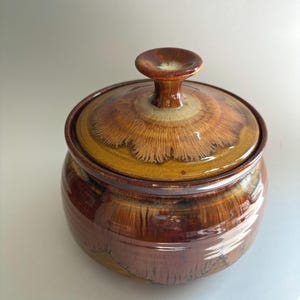 May include: A round ceramic jar with a matching lid featuring a glossy, multi-toned brown glaze. The surface displays a distinctive drip pattern with radiating streaks of lighter tan and deep mahogany, topped with a central knob handle.