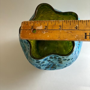 May include: A glass vase with a wavy, scalloped rim and a textured, blue and green mottled exterior. A wooden ruler is placed across the top opening, showing a diameter measurement of approximately 4.25 inches for the vase opening.