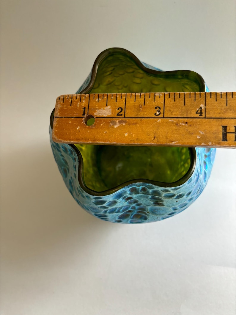 May include: A glass vase with a wavy, scalloped rim and a textured, blue and green mottled exterior. A wooden ruler is placed across the top opening, showing a diameter measurement of approximately 4.25 inches for the vase opening.