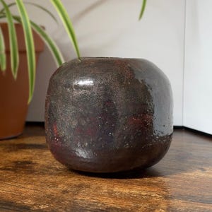 May include: A small, rounded ceramic vase with a dark, mottled finish. The surface features a textured, deep brown and black glaze with subtle hints of red and metallic speckles, creating a rustic, uneven appearance with a glossy sheen.