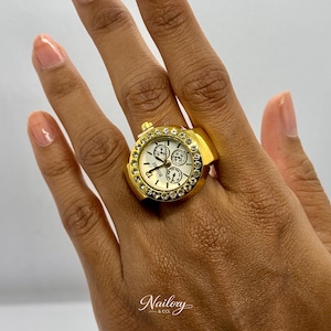May include: A gold-toned ring watch with a white face and three sub-dials. The watch ring is encrusted with clear crystals around the bezel. The ring is on a person's finger.