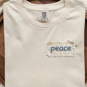 A white t-shirt with the word "Peace" embroidered on it, placed on a wooden tray.