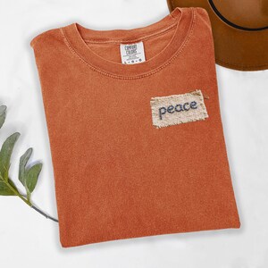 a red t-shirt with a patch on the front that says "peace" and a brown hat placed next to it on a white surface.