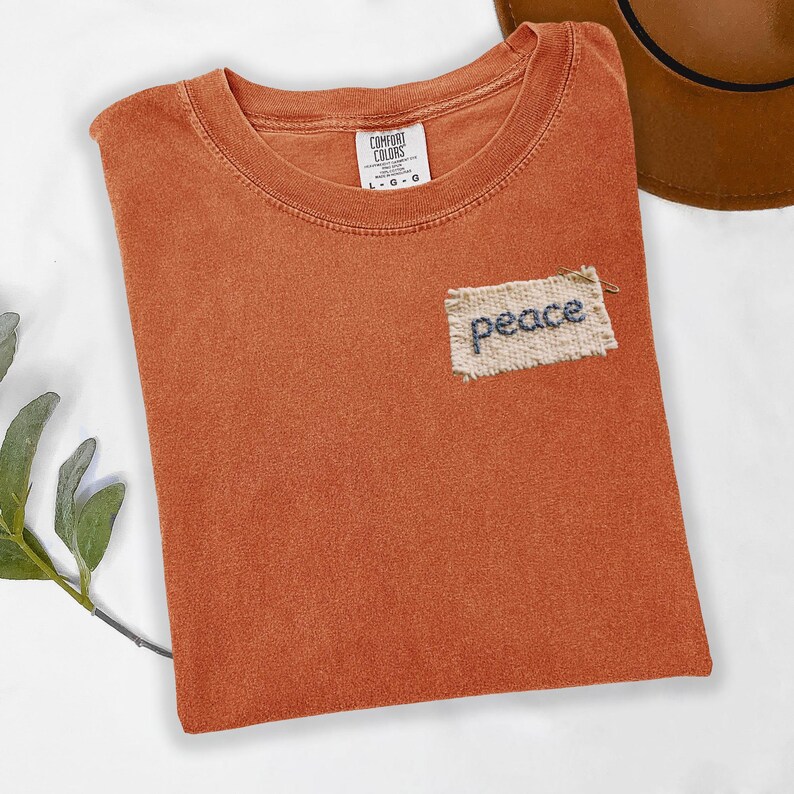 a red t-shirt with a patch on the front that says "peace" and a brown hat placed next to it on a white surface.