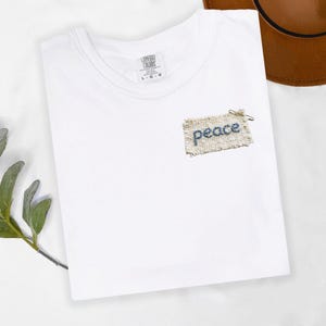 A white t-shirt with a small patch of fabric that says "Peace" on it, placed on a white surface next to a brown hat.
