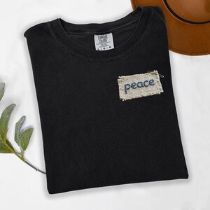 a black t-shirt with a patch on the front that says "Peace" and a brown hat placed next to it on a white surface.
