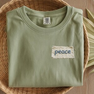 A green t-shirt with a hand-stitched patch that says "peace" is displayed on a woven basket.