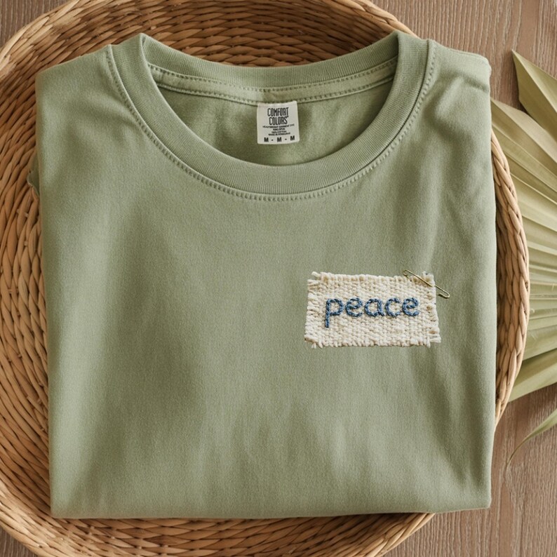 A green t-shirt with a hand-stitched patch that says "peace" is displayed on a woven basket.