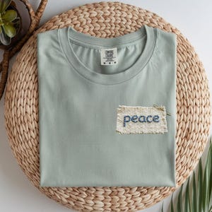 a light green t-shirt with a hand-sewn patch on the front that says "peace". The t-shirt is placed on a woven placemat, and there are some plants visible in the background.