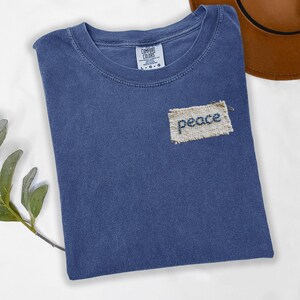 a blue t-shirt with a patch that says "Peace" on it, placed on a white surface next to a brown hat.