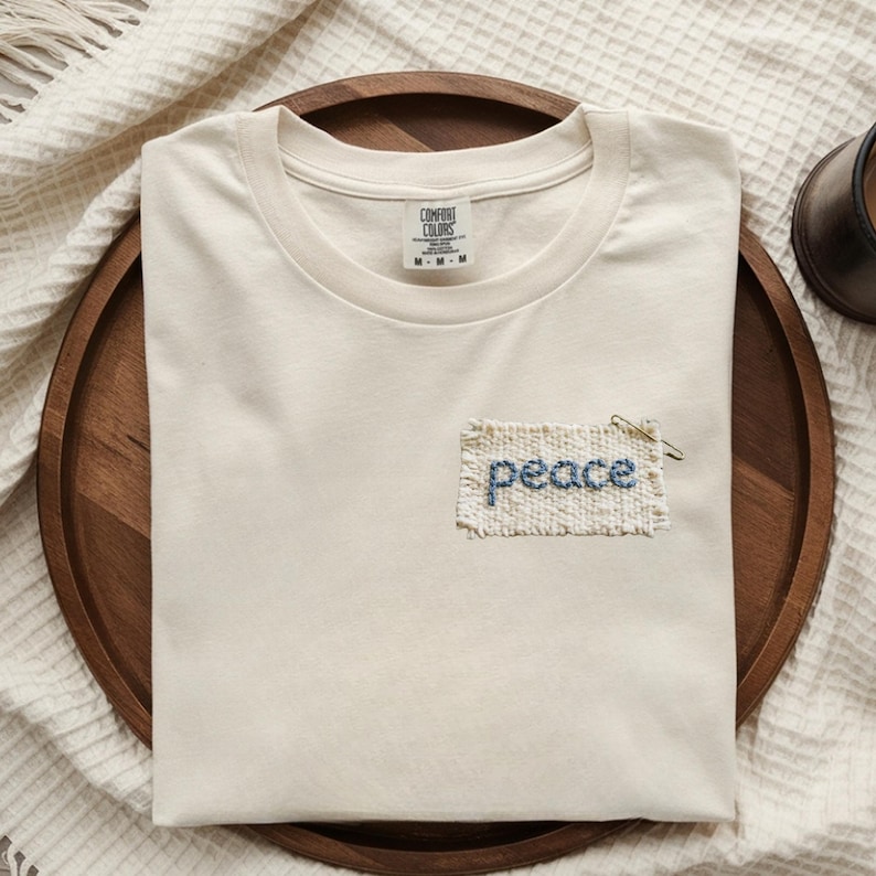 A white t-shirt with the word "Peace" embroidered on it, placed on a wooden tray.