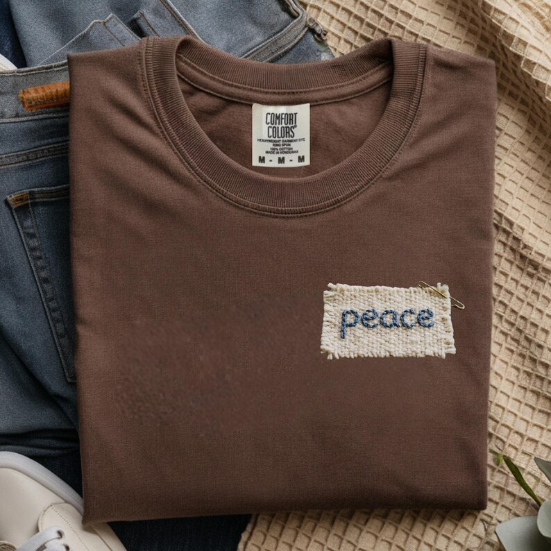 A brown t-shirt with a label that says "Comfortable Colors" and a small patch with the word "Peace" embroidered on it, along with a pair of jeans and a beige blanket.