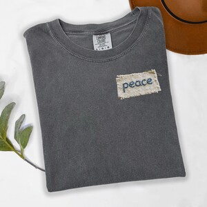 a gray t-shirt with a patch on the front that says "Peace" and a brown hat placed next to it on a white surface.