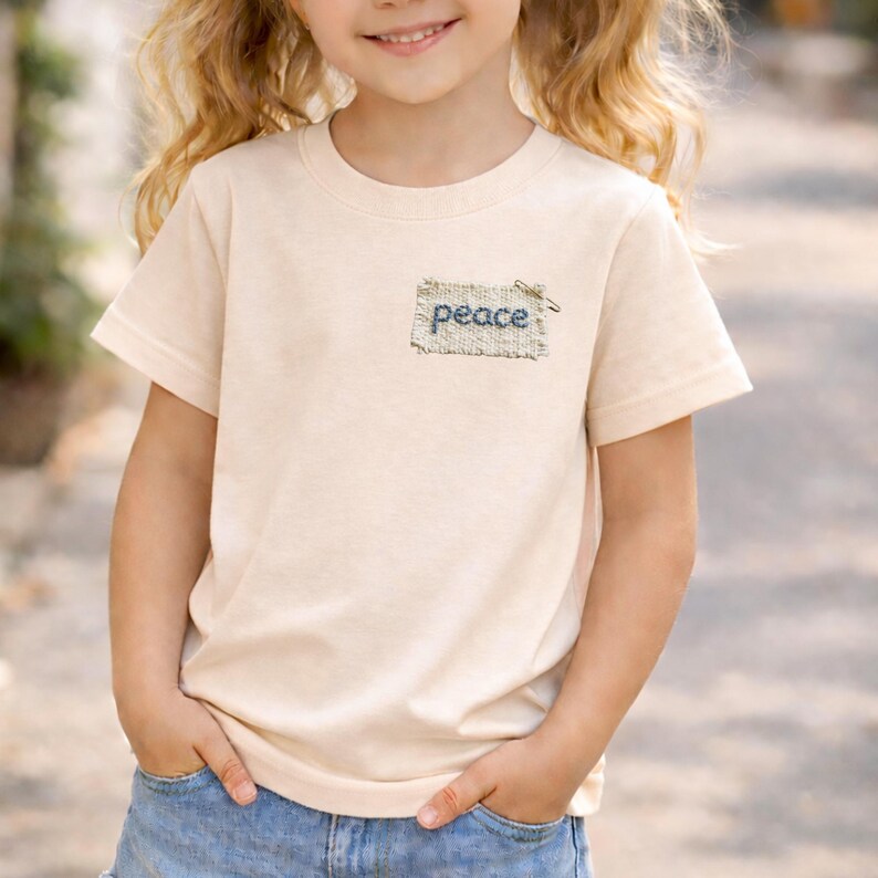 A young girl with blonde hair is smiling and wearing a white t-shirt with the word "Peace" embroidered on it.