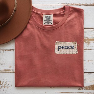 A red t-shirt with a patch that says "Peace" is folded and placed on a white wooden surface, accompanied by a brown hat.