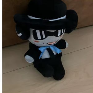 May include: A plush toy character dressed in a black suit, bow tie, and hat. The toy has a light grey face with black-rimmed glasses and a small pink mouth. The plush is sitting on a wooden floor.
