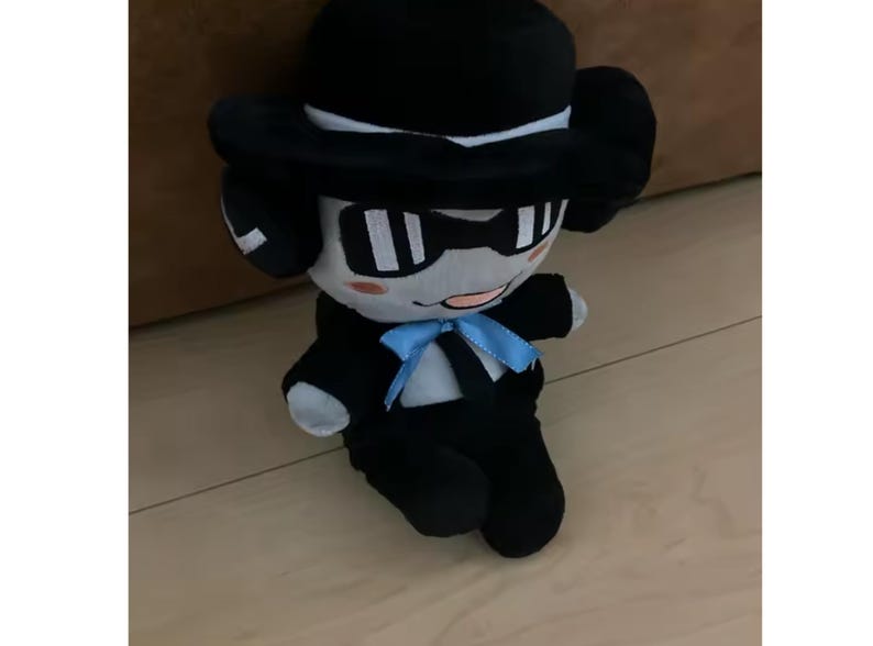 May include: A plush toy character dressed in a black suit, bow tie, and hat. The toy has a light grey face with black-rimmed glasses and a small pink mouth. The plush is sitting on a wooden floor.