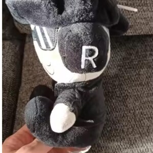 May include: A black and white plush toy of a character wearing a hat and sunglasses, with the letter "R" on its head. The toy has a white face and hands, and is dressed in a black outfit.