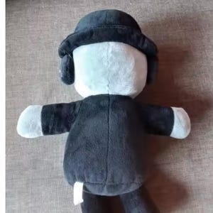 May include: A plush doll with a black coat, hat, and arms, and a white face and hands. The doll has a round hat with ear flaps. The doll's body is black, and the face and hands are white. The doll is sitting on a brown surface.
