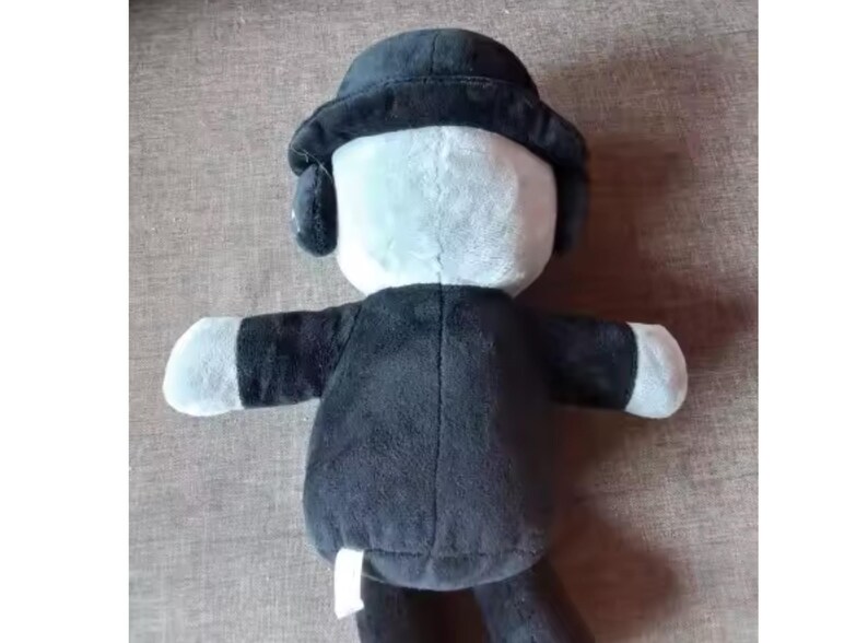 May include: A plush doll with a black coat, hat, and arms, and a white face and hands. The doll has a round hat with ear flaps. The doll's body is black, and the face and hands are white. The doll is sitting on a brown surface.