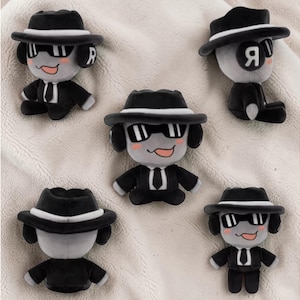 May include: Several plush dog toys dressed in black suits, fedora hats, and sunglasses. The toys are grey and black, with a smiling face and the letter "R" on the side of the head. The toys are arranged on a soft, white surface.