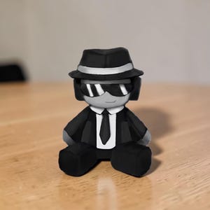 May include: A small figurine of a person in a black suit, hat, and sunglasses. The figure has a white tie and a silver band on the hat. The figurine is sitting on a wooden surface.