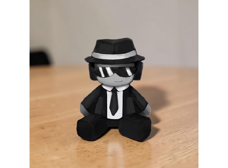 May include: A small figurine of a person in a black suit, hat, and sunglasses. The figure has a white tie and a silver band on the hat. The figurine is sitting on a wooden surface.