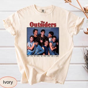 May include: Ivory-colored t-shirt featuring a graphic from the movie "The Outsiders." The design includes a group photo of the cast with the film's title in a red and white font. The shirt is made of a soft material.
