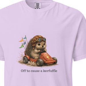 May include: Light purple t-shirt featuring a cartoon hedgehog driving a red go-kart. The hedgehog wears a brown aviator helmet and goggles. The text "Off to cause a kerfuffle" is printed below the image. The shirt has a crew neck and short sleeves.