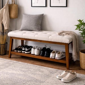 May include: A wooden shoe bench with a cream-colored, tufted cushion and a lower shelf filled with shoes. A gray pillow and a folded blanket rest on the bench. Two framed botanical prints hang on the white wall above the bench. A pair of sneakers sits on the rug.