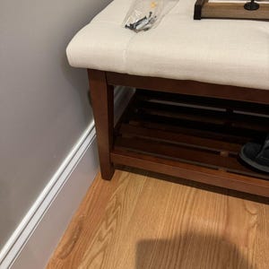 May include: A wooden shoe bench with a cream-colored upholstered seat. The bench has a lower shelf and dark brown legs. A small bag of screws sits on the seat. A wooden tray with a hook is on the bench.