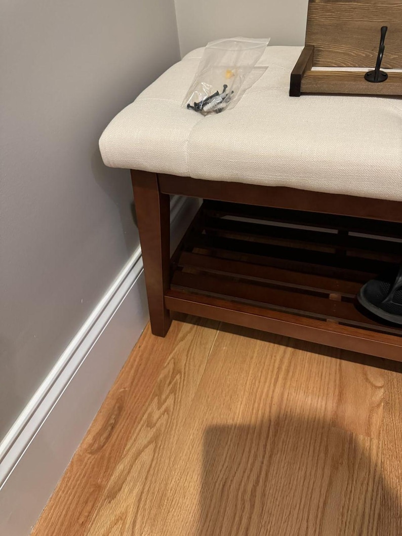 May include: A wooden shoe bench with a cream-colored upholstered seat. The bench has a lower shelf and dark brown legs. A small bag of screws sits on the seat. A wooden tray with a hook is on the bench.