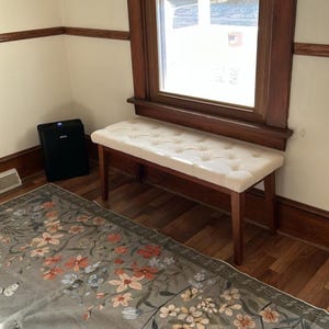 May include: A wooden bench with a cream-colored, button-tufted seat. The bench has four legs and is positioned near a window with a dark wooden frame. The room has light-colored walls and a floral patterned rug.