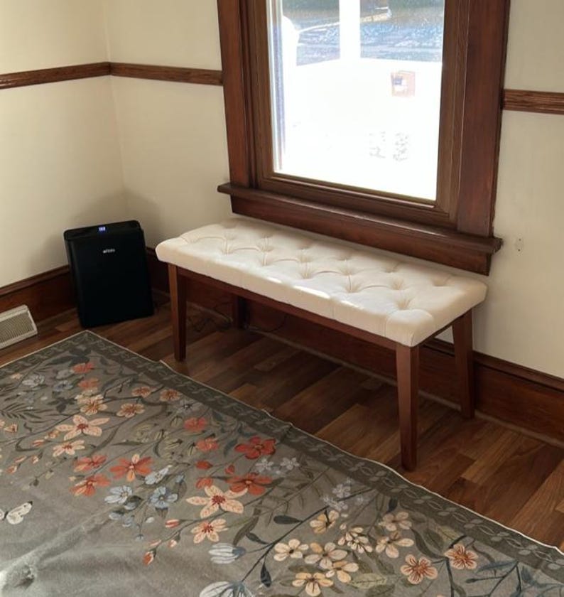 May include: A wooden bench with a cream-colored, button-tufted seat. The bench has four legs and is positioned near a window with a dark wooden frame. The room has light-colored walls and a floral patterned rug.