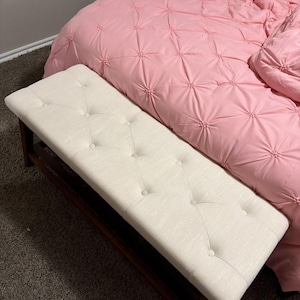 May include: A rectangular, off-white upholstered bench with button tufting. The bench has a dark brown wooden frame. The bench is positioned in front of a pink bedspread with a quilted design.