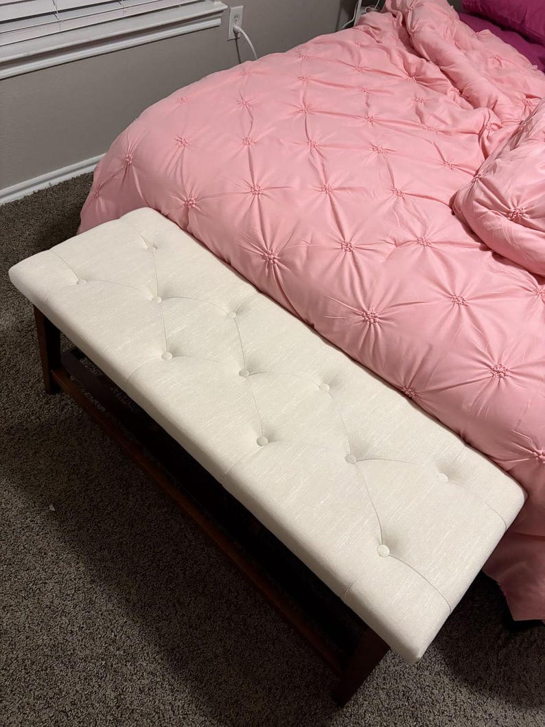 May include: A rectangular, off-white upholstered bench with button tufting. The bench has a dark brown wooden frame. The bench is positioned in front of a pink bedspread with a quilted design.