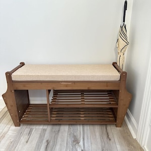 May include: A wooden shoe bench with a cushioned seat and two lower shelves. The bench is made of light brown wood and has a beige cushion. An umbrella with a black handle and a cream and black striped canopy leans against the wall.