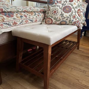 May include: A wooden bench with a light beige, button-tufted seat and a lower shelf. The bench has a dark brown wooden frame. A decorative pillow with a floral pattern rests on the bench. The bench is placed near a bed with a floral patterned bedspread.