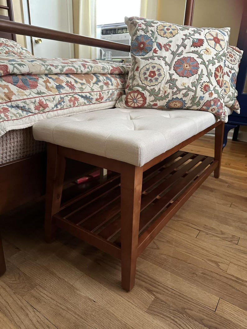 May include: A wooden bench with a light beige, button-tufted seat and a lower shelf. The bench has a dark brown wooden frame. A decorative pillow with a floral pattern rests on the bench. The bench is placed near a bed with a floral patterned bedspread.