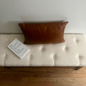 May include: A cream-colored tufted bench with a brown leather pillow and a book titled "101 Essays That Will Change The Way You Think." The bench has a rectangular shape and wooden legs. The pillow is rectangular.