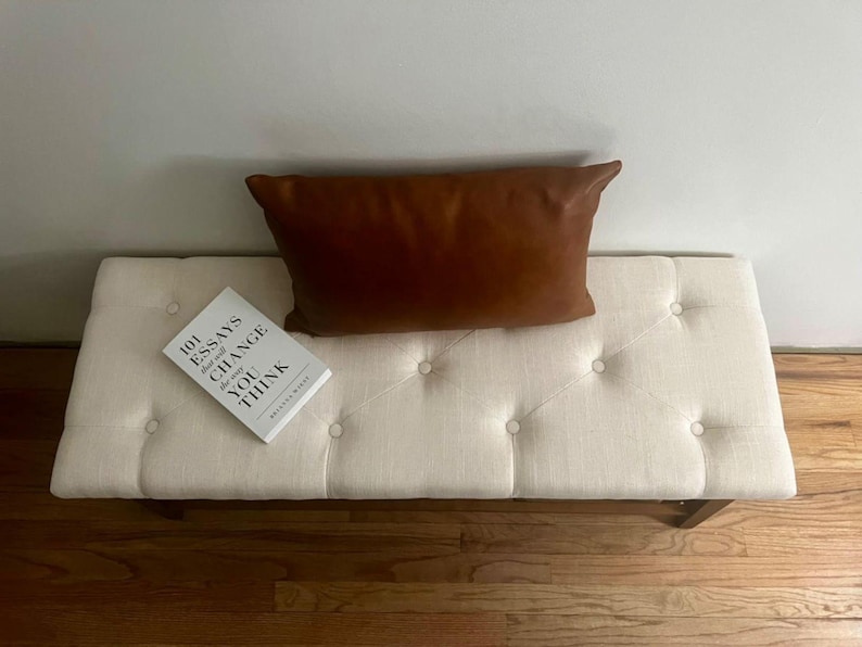 May include: A cream-colored tufted bench with a brown leather pillow and a book titled "101 Essays That Will Change The Way You Think." The bench has a rectangular shape and wooden legs. The pillow is rectangular.