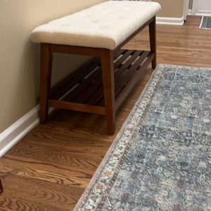 May include: A wooden bench with a cream-colored, button-tufted cushion. The bench has a lower shelf and is set against a beige wall. A patterned rug is in the foreground.