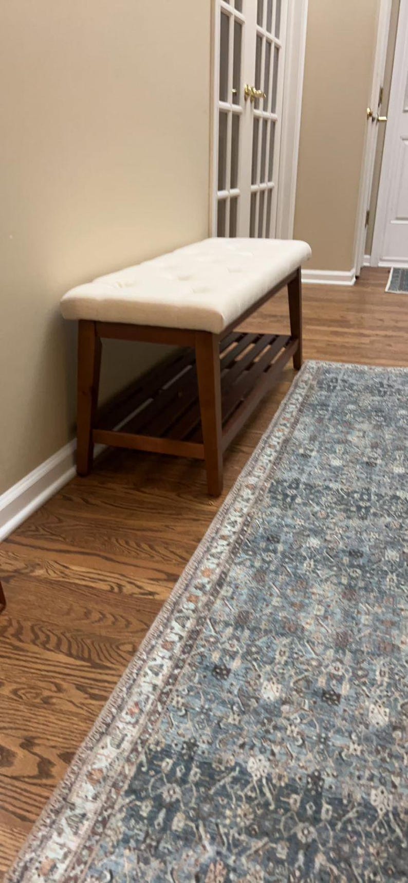 May include: A wooden bench with a cream-colored, button-tufted cushion. The bench has a lower shelf and is set against a beige wall. A patterned rug is in the foreground.