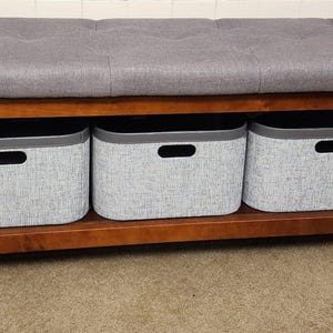 May include: A wooden storage bench with a gray upholstered top and three gray fabric storage bins. The bench has a dark brown frame and is suitable for a bedroom or entryway.