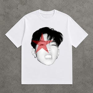 Peut inclure: Un t-shirt blanc à manches courtes arborant un graphique en demi-teintes noir et blanc représentant un visage, avec une grande étoile rouge superposée sur les yeux et le nez. Le motif est centré sur la poitrine du vêtement à col rond.