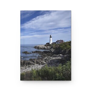 Op de afbeelding: Een notitieboek met een kustlandschap. De omslag toont een witte vuurtoren met een zwart dak, een gebouw met een rood dak en een rotsachtige kustlijn onder een blauwe lucht met wolken. Het notitieboek is rechthoekig en verticaal.