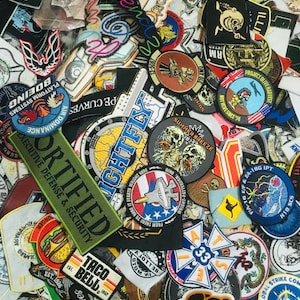 Op de afbeelding: Een verzameling geborduurde patches met diverse ontwerpen en tekst. Patches omvatten militaire, luchtvaart- en commerciële thema's, met tekst zoals "Certified Executive Defense & Security" en "Taco Bell". De patches zijn in verschillende vormen en kleuren.
