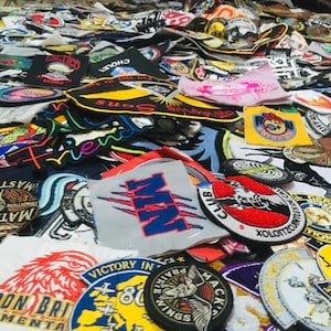 May include: A collection of embroidered patches with various designs and text. Patches feature eagles, wings, text like "Victory In", "Mackinaw Wings", and "Sons". Colors include red, blue, yellow, and black. The patches are scattered across a surface.