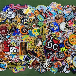 May include: A large collection of assorted embroidered patches featuring various colorful designs, including sports team logos, cartoon characters, food items, text graphics, and cultural symbols, all arranged in a dense, overlapping pile.